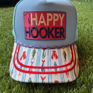 May include: A light blue trucker hat featuring a rectangular patch with the text HAPPY HOOKER in dark blue and beige, accompanied by a fishhook graphic. The brim displays a white background with a repeating pattern of colorful fish and lobsters.