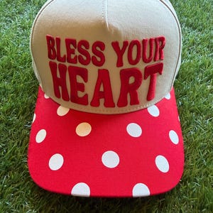 Puede incluir: Una gorra de béisbol beige con el texto bordado en rojo BLESS YOUR HEART en el panel frontal. La gorra tiene un botón rojo en la parte superior y una visera plana decorada con un patrón de lunares blancos sobre un fondo de tela roja.