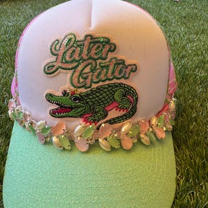 Puede incluir: Una gorra tipo trucker blanca con visera verde menta y malla trasera rosa. El frente presenta un parche bordado con el texto "Later Gator" sobre un caimán verde con corona. Una hilera de pedrería rosa y verde menta bordea la base de la gorra.