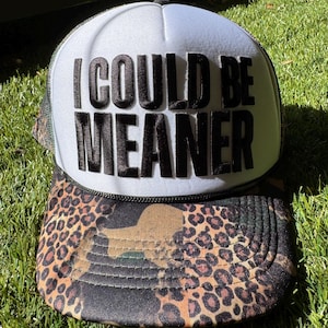 Op de afbeelding: Truckerpet met een wit voorpaneel en de zwarte tekst "I COULD BE MEANER". De klep heeft een luipaardprint. De pet heeft een zwarte mesh achterkant en een zwart touwdetail.
