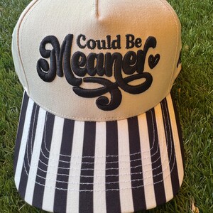 Pu&ograve; includere: Un cappellino da baseball beige con visiera a righe bianche e nere. Il cappellino presenta la frase ricamata "Could Be Meaner" in corsivo nero. Il cappellino &egrave; appoggiato su uno sfondo di erba verde.