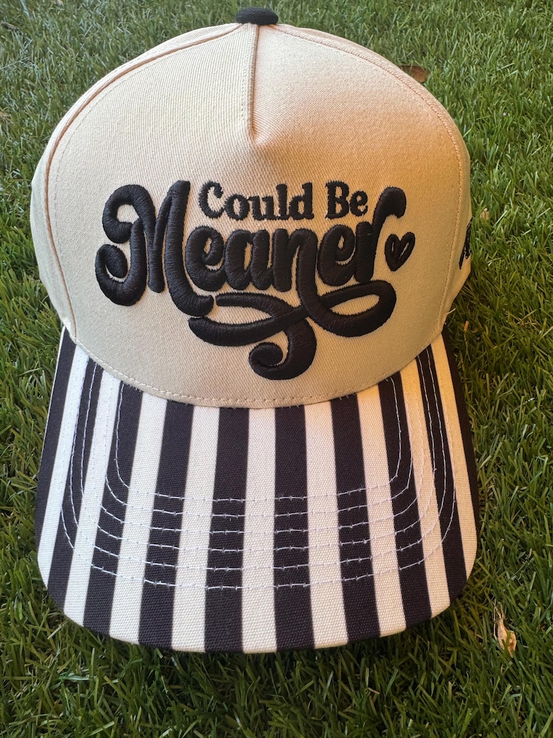 Pu&ograve; includere: Un cappellino da baseball beige con visiera a righe bianche e nere. Il cappellino presenta la frase ricamata "Could Be Meaner" in corsivo nero. Il cappellino &egrave; appoggiato su uno sfondo di erba verde.
