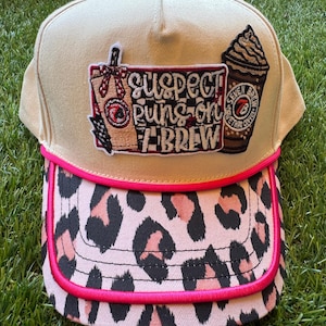 May include: Tan baseball cap with a pink leopard print brim and a pink trim. The cap features embroidered patches with coffee-themed graphics and the text "Suspect Puns-on Brew" and "Seven Brew Coffee".