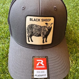 Puede incluir: Una gorra tipo trucker de color gris carbón con malla negra en la parte posterior y un parche cuadrado al frente que muestra una ilustración negra de una oveja y el texto BLACK SHEEP. Una etiqueta rectangular en la visera muestra el logo Richardson.