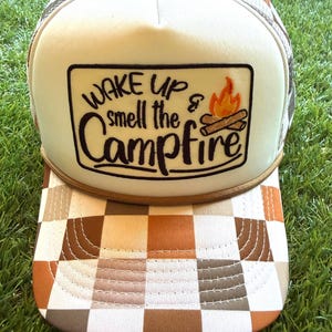 Può includere: Un cappellino trucker con pannello frontale bianco e una toppa rettangolare che riporta la scritta "WAKE UP & smell the Campfire" in nero, insieme a un ricamo di un falò. La visiera e i pannelli posteriori presentano un motivo a scacchi marrone, beige e bianco.