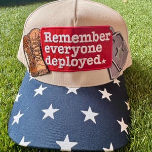 May include: A beige baseball cap with a navy blue brim featuring a white star pattern. The front panel displays a rectangular red patch with the white embroidered text "Remember everyone deployed" flanked by a brown boot and a silver dog tag graphic.