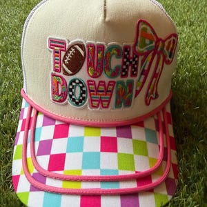 Op de afbeelding: Een witte truckerpet met een kleurrijke geblokte klep en roze bies. Het voorpaneel is voorzien van een geborduurde patch met de tekst TOUCH DOWN, een bruin football-icoon en een grote, veelkleurige strik aan de rechterkant.