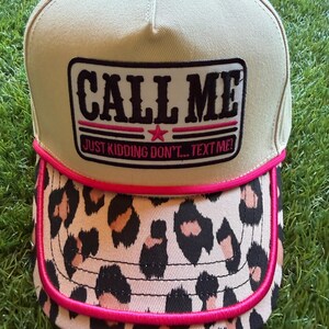 May include: A beige baseball cap featuring a rectangular patch with the black text CALL ME and the pink text JUST KIDDING DON'T... TEXT ME! The cap has a leopard print brim and a bright pink cord trim separating the crown from the brim.