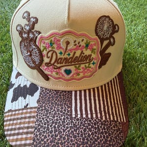 May include: A beige baseball cap featuring a central embroidered patch with the word Dandelion in script, surrounded by floral and dandelion seed head motifs. The brim displays a patchwork pattern of cow print, leopard print, and stripes.