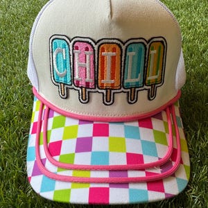 May include: A white trucker hat featuring an embroidered patch of five colorful popsicle icons with the text CHILL spelled out across them. The brim displays a vibrant multicolored checkerboard pattern with pink piping trim along the edges.