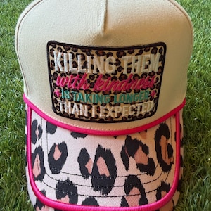 May include: A beige baseball cap featuring a leopard print patch with the embroidered text "KILLING THEM with kindness IS TAKING LONGER THAN I EXPECTED" in white, pink, and teal. The brim has a leopard print pattern and bright pink piping.
