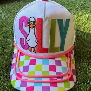 Puede incluir: Una gorra tipo camionero blanca con visera de cuadros multicolores y un panel frontal con la palabra SILLY en letras grandes de tela de colores. La letra I está sustituida por un pájaro blanco bordado con gafas de sol verdes.