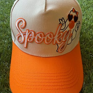 May include: A beige baseball cap with an orange brim and an orange button on top. The front features the word Spooky embroidered in orange script with a white outline, alongside an embroidered white ghost wearing heart-shaped sunglasses.