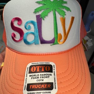 May include: A trucker hat with a white front panel and an orange brim. The word "Sally" is embroidered in pink, blue, purple, and orange, with a green palm tree. The hat has a mesh back and a snap closure. The hat has a tag that says "OTTO TRUCKER".