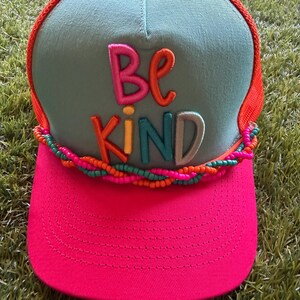 Pode incluir: Um boné trucker com painel frontal azul claro, aba rosa brilhante e parte traseira de rede laranja. A frente apresenta o texto bordado "BE KIND" em letras multicoloridas. Um acabamento trançado de pequenas contas rosas, laranjas e turquesas situa-se acima da aba.
