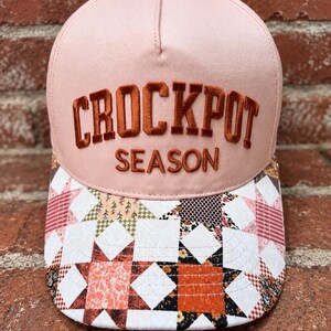 Può includere: Un cappellino da baseball rosa chiaro con un bottone superiore giallo. La parte anteriore presenta la scritta ricamata "CROCKPOT SEASON" in arancione scuro. La visiera mostra un motivo patchwork multicolore con quadrati bianchi, arancioni e floreali.
