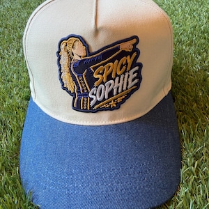 May include: A beige baseball cap with a blue denim brim and a top button. The front features an embroidered patch depicting a person with blonde hair pointing, accompanied by the text SPICY SOPHIE in bold yellow and white lettering.