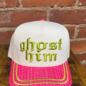 May include: A white baseball cap featuring the embroidered text "ghost him" in lime green on the front panel. The cap has a bright pink brim with a subtle grid pattern and lime green piping accents, topped with a small pink button at the crown.