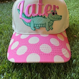 Puede incluir: Gorra de béisbol beige con visera rosa con lunares blancos. La gorra presenta la palabra "Later" en rosa sobre un caimán verde con gafas de sol.
