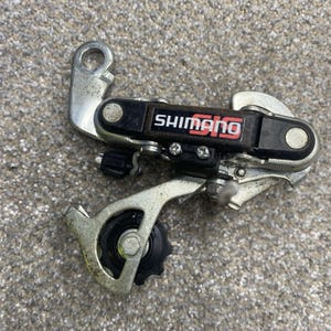 May include: A silver and black Shimano SIS bicycle rear derailleur featuring a metal mounting bracket, a black plastic housing with white and red text, and a black pulley wheel. The component includes adjustment screws and a cable tension barrel.
