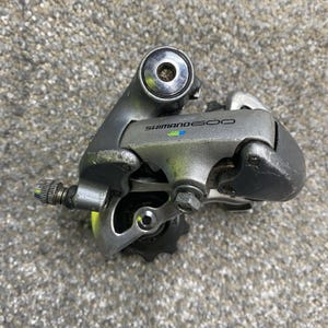 Può includere: Un deragliatore posteriore per bicicletta Shimano 600 color argento con finitura metallica, pulegge nere e un registro di tensione del cavo. Il componente presenta il logo Shimano 600 in nero con piccoli accenti verdi e blu sul corpo.