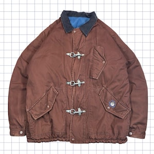 May include: A brown jacket with a blue collar and lining. The jacket has three toggle closures, two front pockets, and a small embroidered patch on the right pocket. The jacket has a drawstring at the bottom and is made of a durable material.