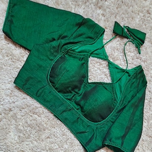 May include: A dark emerald green blouse with a square neckline and long sleeves. The blouse has a satin finish and a tie closure at the back. The garment is displayed on a neutral, textured surface.