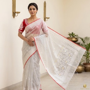 May include: A white sheer saree featuring intricate white floral embroidery and a distinct red border. The garment is paired with a short-sleeved red blouse that has matching white embroidery along the sleeves and neckline, creating a cohesive look.