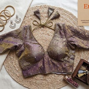 May include: A textured tissue silk blouse in shades of purple and gold with a deep V-neckline, short sleeves, and back tie closures. The blouse features small white embroidered accents and is displayed with four gold bangles and two earrings.
