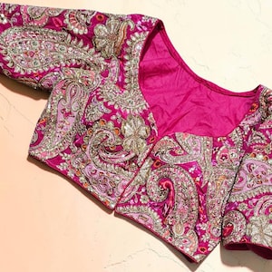 May include: A vibrant magenta blouse featuring intricate silver and gold metallic embroidery in a paisley pattern. The garment has short sleeves, a wide scoop neckline, and a solid magenta interior lining, showcasing detailed needlework throughout.