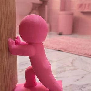 May include: A vibrant pink door stop in the form of a person, appearing to push against a door. The door stop is made of a solid material and is placed against a wooden door. The background shows a pink bathroom.