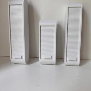 May include: Three white rectangular storage containers with rounded top edges and vertical sliding tambour doors. Each container features a small horizontal tab at the bottom of the door for opening and closing the unit, presented in three different heights.