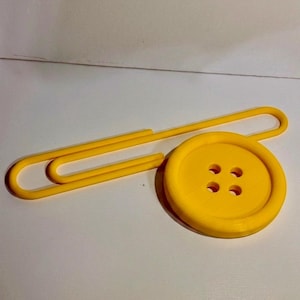 May include: A bright yellow oversized paperclip attached to a large, round, four-hole yellow button. The object is constructed from a solid, smooth material with a uniform matte finish, featuring a classic paperclip shape and a circular button design.
