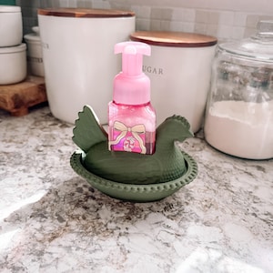May include: A pink soap dispenser with a bow design sits in a green ceramic hen-shaped holder. The dispenser is on a countertop with white canisters and a glass jar in the background. The hen holder is approximately 6 inches long.