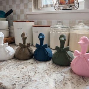 May include: A collection of decorative goose figurines in various colors, including white, brown, blue, green, and pink. Each goose has a bow around its neck. The figurines are displayed on a countertop near kitchen canisters.