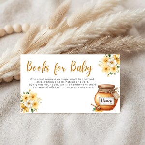 Pode incluir: Cartão de convite para chá de bebé com o texto "Books for Baby" em escrita dourada. O cartão apresenta ilustrações em aquarela de flores amarelas e um pote de mel. O texto pede aos convidados que tragam um livro em vez de um cartão.