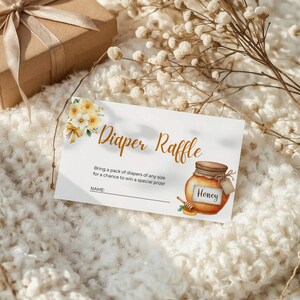 Puede incluir: Una tarjeta de invitación para baby shower con las palabras "Diaper Raffle" en escritura elegante. La tarjeta presenta una ilustración de acuarela de un tarro de miel y flores. Una caja de regalo envuelta está en el fondo.