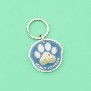 May include: A round keychain with a blue paw print design and the words "BORN TO ROAM." The paw print has white pads and a mountain scene with a red sun. The keychain has a silver ring.