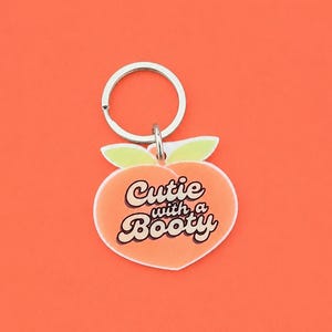 May include: A peach-shaped keychain with the phrase "Cutie with a Booty" in white and brown lettering. The keychain has a silver ring and a light orange background.
