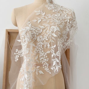 May include: White floral lace fabric with beaded embellishments. The lace features a delicate floral design with small, white beads and sequins. The fabric is sheer and lightweight, making it ideal for wedding dresses, bridal accessories, and other special occasion garments.