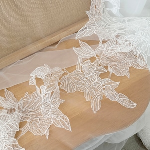 May include: White lace trim with a floral design on a sheer white fabric. The lace is delicate and intricate, with a repeating pattern of flowers and leaves.