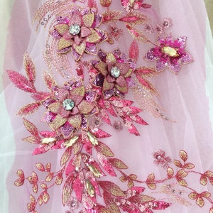 Könnte beinhalten: Gestickte Blumenapplikation mit rosa und goldenen Pailletten, Perlen und Kristallen. Das Design zeigt mehrere Blumen und Blätter auf einem transparenten rosa Stoff. Die Applikation eignet sich zum Verzieren von Kleidungsstücken oder Accessoires.