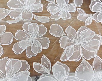 May include: Close-up of white floral lace fabric. The lace features large, detailed flower appliques with delicate beaded centers and outlines. The flowers are arranged across a sheer, net-like background, creating a textured and elegant design.