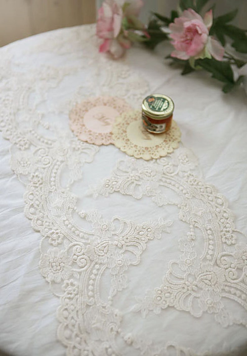 Puede incluir: Borde de encaje blanco con un delicado patr&oacute;n floral. El encaje est&aacute; sobre un mantel blanco con dos manteles individuales y un peque&ntilde;o tarro de miel.