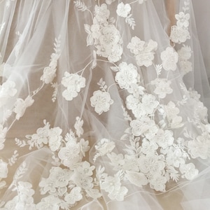 May include: White floral lace fabric with a delicate tulle background. The lace features 3D flowers with pearl embellishments.