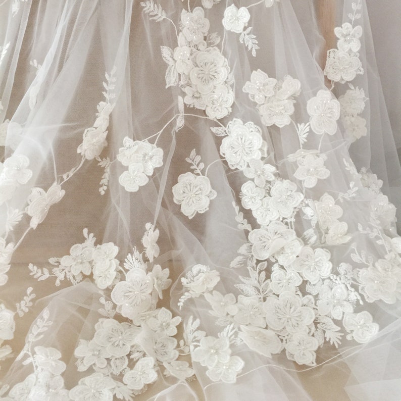 May include: White floral lace fabric with a delicate tulle background. The lace features 3D flowers with pearl embellishments.