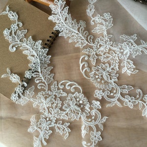 以下が含まれることがあります： 花柄の白いレースアップリケ2つ。アップリケは繊細で複雑で、小さな花と葉がデザインされています