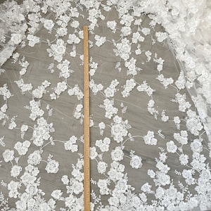 May include: White floral lace fabric with a ruler for scale. The lace has a delicate, intricate design with small, raised flowers and leaves. The fabric is sheer and has a soft, flowing texture.