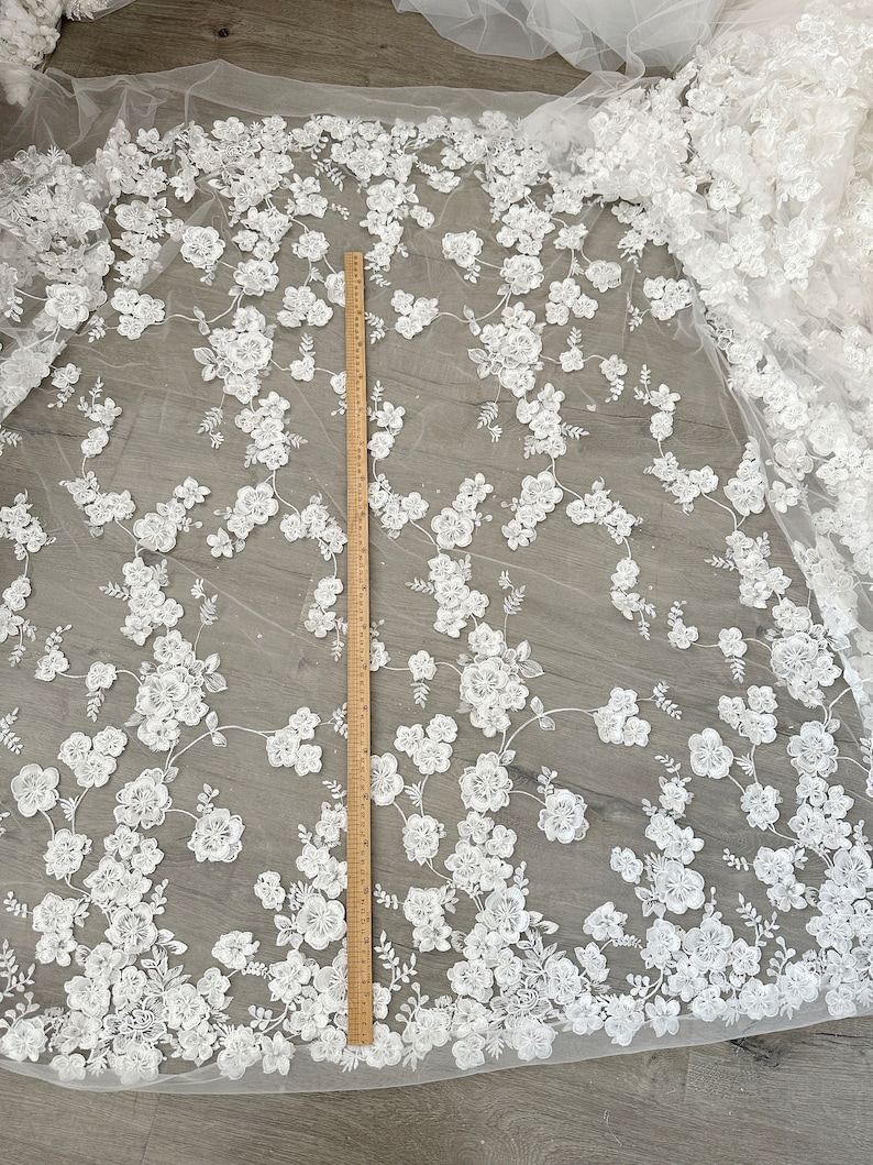 May include: White floral lace fabric with a ruler for scale. The lace has a delicate, intricate design with small, raised flowers and leaves. The fabric is sheer and has a soft, flowing texture.