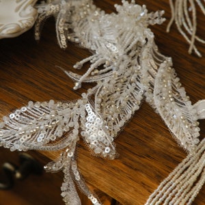 May include: Close-up of white beaded lace trim with sequins and small beads. The trim is arranged in a leaf-like pattern, with delicate details and a textured appearance. The lace is on a wooden surface.